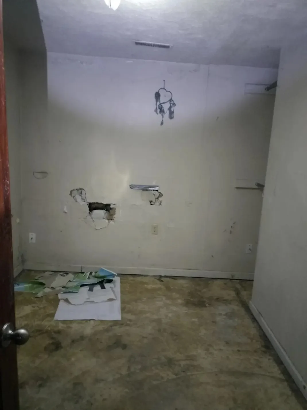 One room before renovation, with damaged walls, removed fixtures, exposed openings, and stripped flooring.